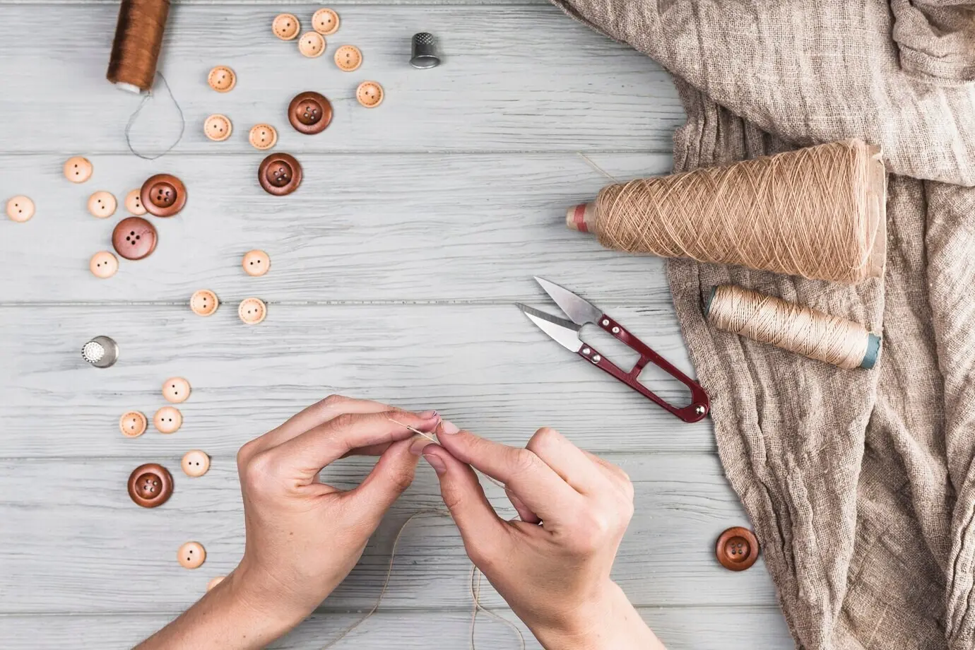 Nahaufnahme einer Frauenhand, die einen Faden in eine Nadel einfädelt; mit Knopf, Cutter und Stoff vor hölzernem Hintergrund.